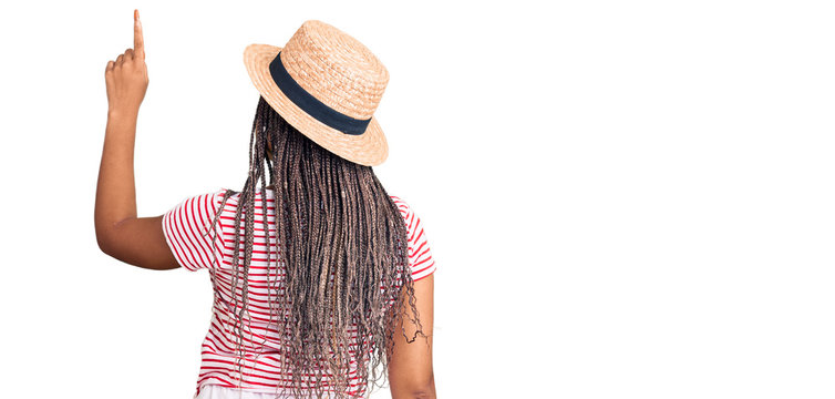 Young african american woman with braids wearing summer hat posing backwards pointing ahead with finger hand