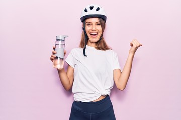Young beautiful girl wearing bike helmet drinking bottle of water pointing thumb up to the side smiling happy with open mouth