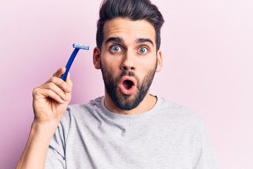 Young handsome man with beard holding razor scared and amazed with open mouth for surprise,...