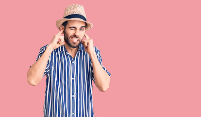Young handsome man with beard wearing summer hat and striped shirt covering ears with fingers with annoyed expression for the noise of loud music. deaf concept.