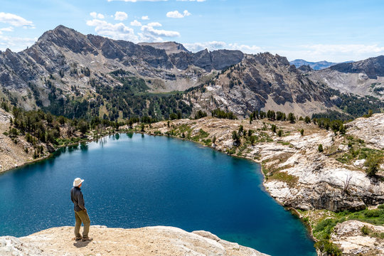 Man, hiker, standing, watching a scene of an alpine lake and rugged mountains with trees, rocks, and puffy clouds in the blue sky, Liberty Lake, Ruby Mountain Range, Elko, Nevada