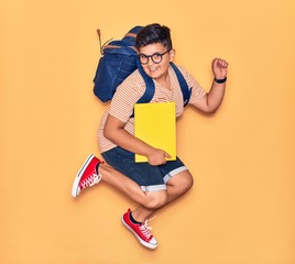 Adorable student boy wearing glasses and backpack smiling happy. Jumping with smile on face holding book celebrating with fist up over isolated yellow background.