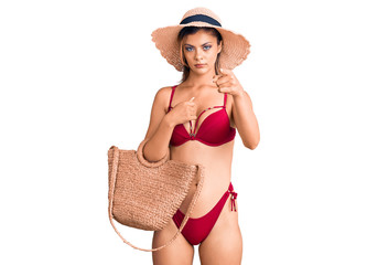 Young beautiful woman wearing bikini and hat holding summer wicker handbag pointing with finger to the camera and to you, confident gesture looking serious