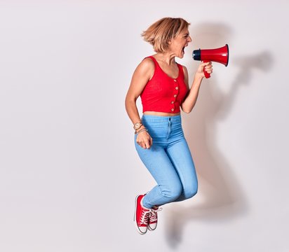 Young beautiful caucasian woman screaming using megaphone. Jumping over isolated white background