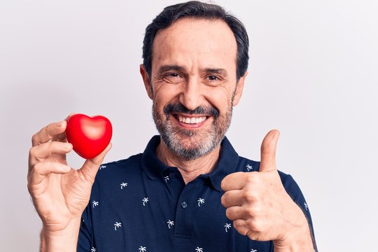 Middle age handsome romantic man holding plastic heart over isolated white background smiling happy and positive, thumb up doing excellent and approval sign