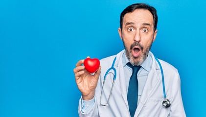 Middle age handsome cardiologist man wearing stethoscope holding plastic heart scared and amazed...