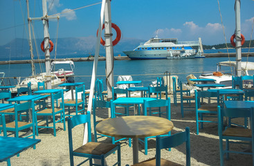 Greek traditional restaurant with turquoise empty chairs and a ship in the background
