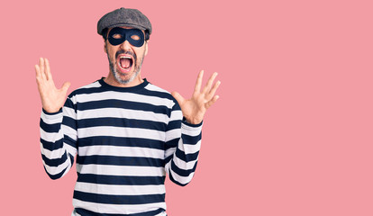 Middle age handsome man wearing burglar mask celebrating mad and crazy for success with arms raised and closed eyes screaming excited. winner concept