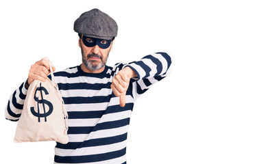 Middle age handsome man wearing burglar mask holding money bag with angry face, negative sign showing dislike with thumbs down, rejection concept