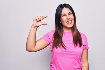 Obraz na płótnie Canvas Young beautiful brunette woman wearing casual pink t-shirt standing over white background smiling and confident gesturing with hand doing small size sign with fingers looking and the camera. Measure