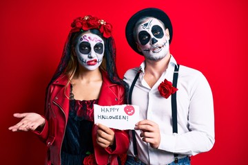 Couple wearing day of the dead costume holding happy halloween paper clueless and confused expression. doubt concept.