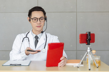 Young asian doctor wear eyeglass headset and stethoscope talking through system online to patient. Using tablet and smartphone. New normal life style.