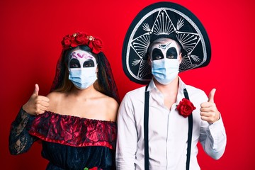 Couple wearing day of the dead costume wearing medical mask smiling happy and positive, thumb up doing excellent and approval sign