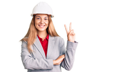 Beautiful young woman wearing architect hardhat smiling with happy face winking at the camera doing victory sign. number two.
