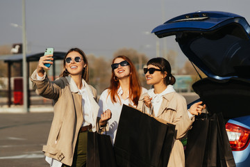 Obraz premium Young women at the car with shopping bags. Girls take selfie