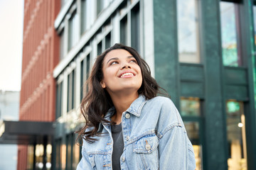 Confident happy beautiful young hipster African American woman wearing denim jacket looking up standing on city street outdoors dreaming, thinking or good future on urban buildings background. - obrazy, fototapety, plakaty