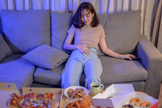 Full After Eating Fast Food When Takeout And Delivery At Night. Takeaway Back Home And Watching TV. Asian Woman Lifestyle In Living Room. Social Distancing And New Normal.