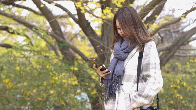 4K Happy Asian woman tourist using smartphone camera to selfie with beautiful yellow ginkgo biloba leaves falling down during autumn at the park in Japan. Japan Travel and season change concept.