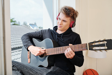 Smiling creative teenager in headphones sitting on window sill and playing guitar