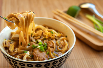 In the foreground a wide yellow noodle on a fork. In the background, a plate of Asian noodle soup with dark broth, chunks of beef and greens in a deep round plate. The plate is on a bamboo tablecloth.