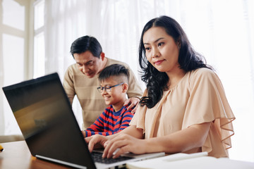 Concentrated Asian woman working on laptop when her husband helping preteen son with homework