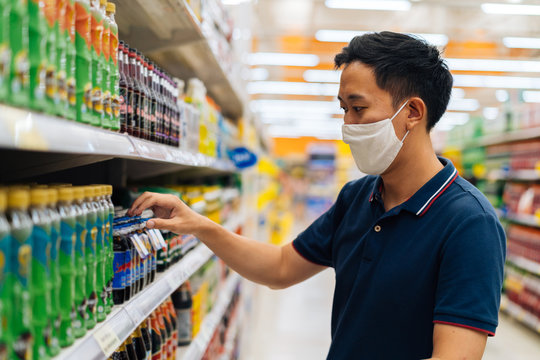 Young Adult Asian Man Wearing A Face Mask While Shopping With Cart Trolley In Grocery Supermarket Store. He's Choosing To Buy Products In The Grocery Store During Covid 19 Crisis In Thailand