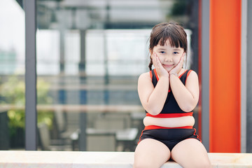 Portrait of smiling little Asian girl sitting on edge of swimming pool, touching cheeks and looking at camera