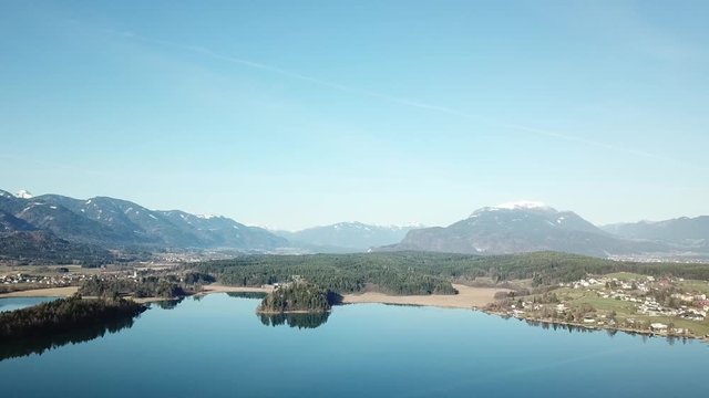 A drone flight above the Faaker See in Austria. There are high Alpine ranges around, reflecting in the crystal clear and calm surface of the lake. Small villages around the lake. Snow capped mountains