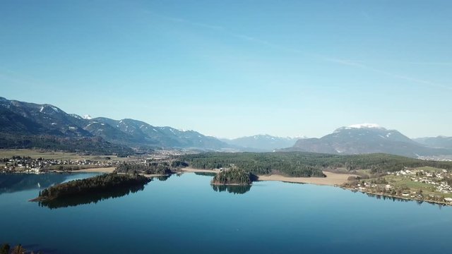 A drone flight above the Faaker See in Austria. There are high Alpine ranges around, reflecting in the crystal clear and calm surface of the lake. Small villages around the lake. Snow capped mountains
