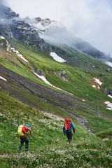 Fototapeta na wymiar Back view of two man with backpacks hiking together in mountains. Male hikers walking down mountain path in grassy hillside meadow. Concept of traveling, hiking and active leisure.