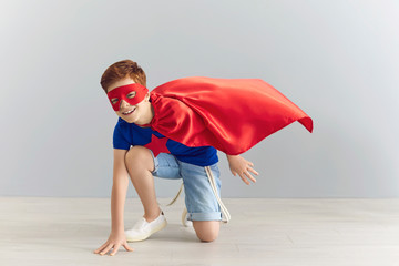 Cheerful boy in superhero costume ready to fight evil on grey background. Positive child pretending...