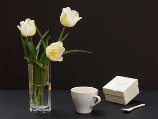 Bouquet of yellow tulips in vase with gift box and cup on a black background.