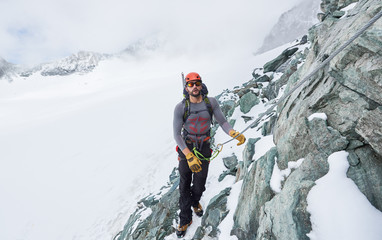Male alpinist in sunglasses and safety helmet holding fixed rope while climbing snowy mountain. Mountaineer ascending natural rock formation. Concept of winter rock climbing.