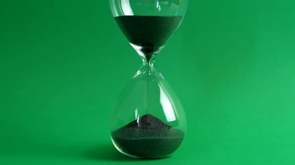 Shot of a sand clock measuring time while the sand is falling down against the green background - old classic timer. Extreme close up of a transparent hourglass with flowing black sand - time concept.