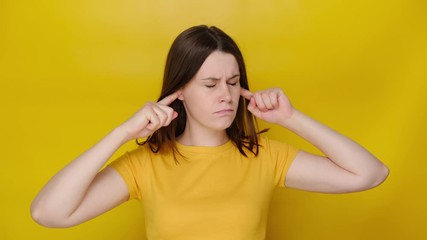 Annoyed irritated young woman covering ears and gesturing No, avoiding advice, ignoring unpleasant noise, loud voices, wears t-shirt, isolated on yellow studio background. Don't want to listen!