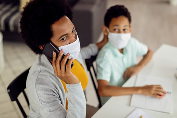 African American mother talking on the phone while homeschooling her son during COVID-19 epidemic.