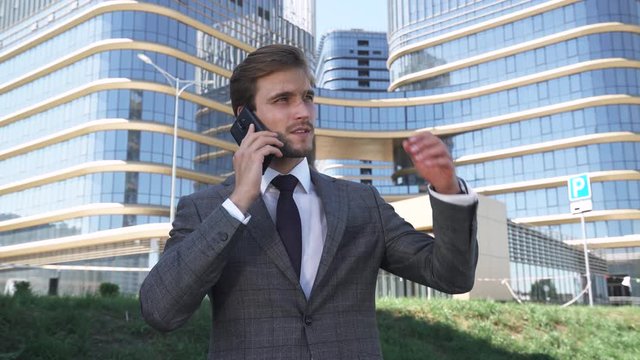 Young businessman in a suit standing near a glass skyscraper and talking on a mobile phone, top manager near the modern office.