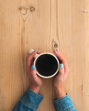 Female hands holding onto a white cup filled with black Americano coffee.  Lady is wearing faded denim shirt and hands are rested on pale natural wood grain surface or table.  Flat lay style image