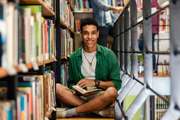Young male student study in the library reading book while sitting near bookshelf and looking at...