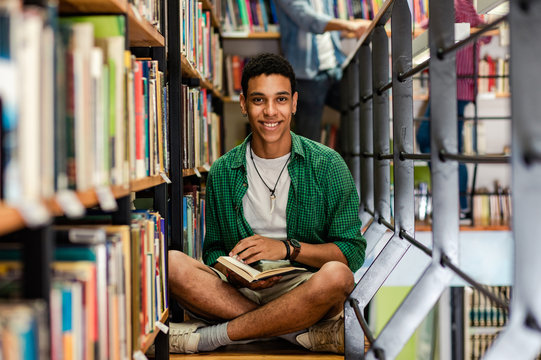 Young male student study in the library reading book while sitting near bookshelf and looking at camera,
