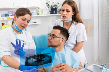 Female dentist with an assistant showing dental x-ray of teeth to a male patient in the clinic.