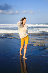 Pretty young woman walking on black sand beach. Caucasian woman wearing yellow sportswear and white blouse. Happiness and freedom. Travel lifestyle. Copy space.