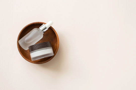 Serum in glass bottle with pipette and facial cream moisturizer in wooden plate on light beige background with copy space, top view. Minimalist set natural cosmetic beauty products for skin care