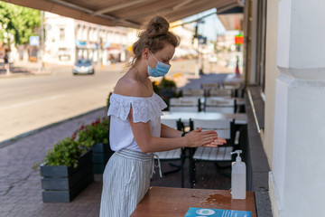 Kyiv (Kiev), Ukraine - August 08, 2020: A beautiful young woman in a mask on the street is disinfecting hands with antiseptic near the restaurant or a cafe on a sunny summer day