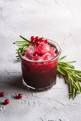 Fresh ice cold fruit cocktail in glass, refreshing summer red currant berry drink with rosemary leaf on stone concrete background, angle view