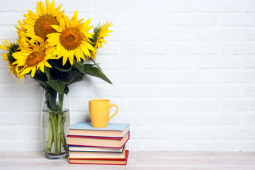 A bouquet of sunflowers in a vase with Stack of books and yellow mug against the white brick wall.