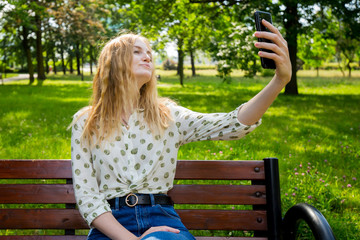 Beautiful young woman with blonde hair taking selfie at the city park. Girl sitting on a bench in a sunny day. Active life concept