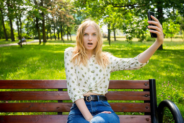 Beautiful young woman with blonde hair taking selfie at the city park. Girl sitting on a bench in a sunny day. Active life concept