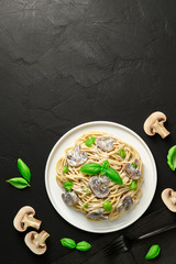 Spaghetti with creamy sauce, mushrooms and green basil on a dark background. Italian pasta with champignons in a gray plate. Flat lay with place for text, vertical orientation.