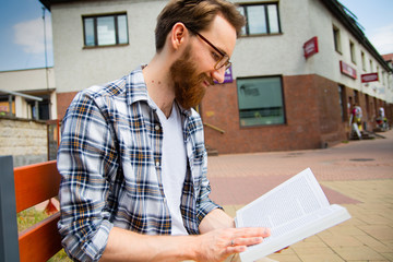 Young adult man with a beard sitting on a city bench and reading a book.